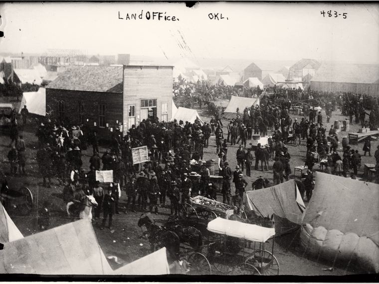 A crowd outside a frame land office building, LANdOFFice. OKL. handwritten across the sky. Library of Congress ggbain.02285.