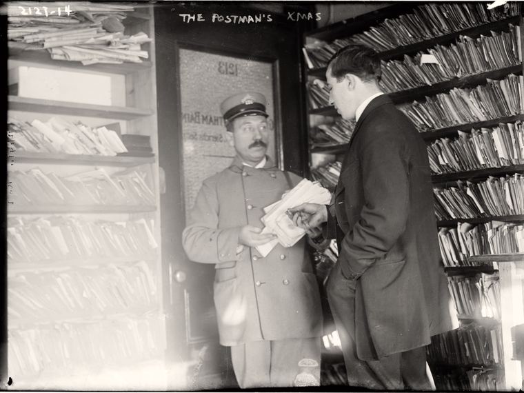 A postman with an armful of mail among stacked parcels, The Postman's Xmas handwritten at the top. Library of Congress ggbain.08872.